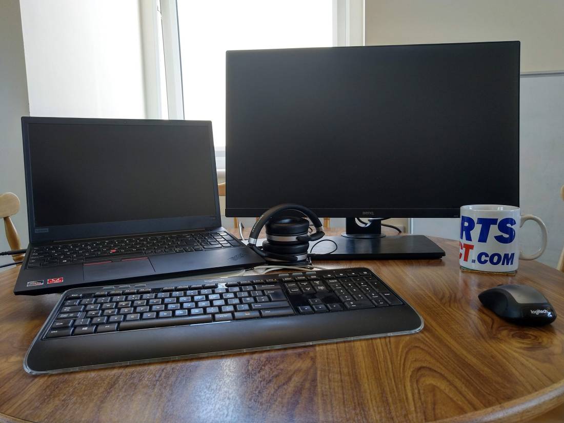 A kitchen table with a laptop on the left and a large external screen on the right. A keyboard and mouse are visible in the foreground, with a large Sports Direct mug on the right and a pair of over-ear headphones charging in the centre
